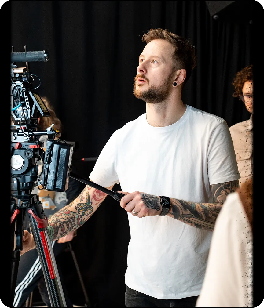 Tattooed filmmaker in a white t-shirt adjusting a professional video camera on a tripod in a dimly lit studio.