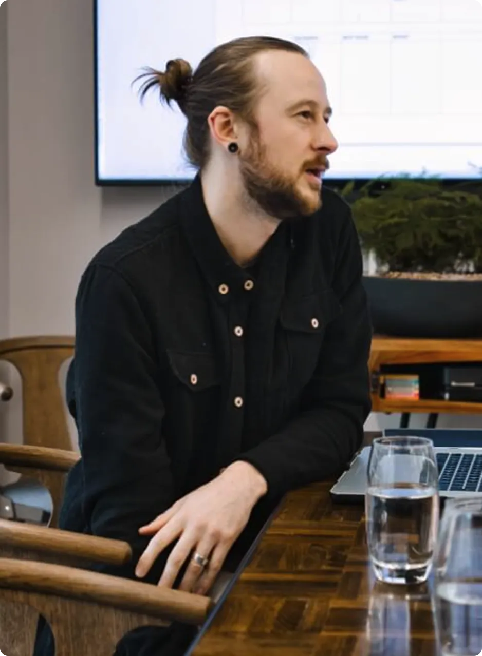 Man with a beard and tied-back hair sitting at a wooden table, speaking during a meeting with a laptop and glasses of water in front of him.