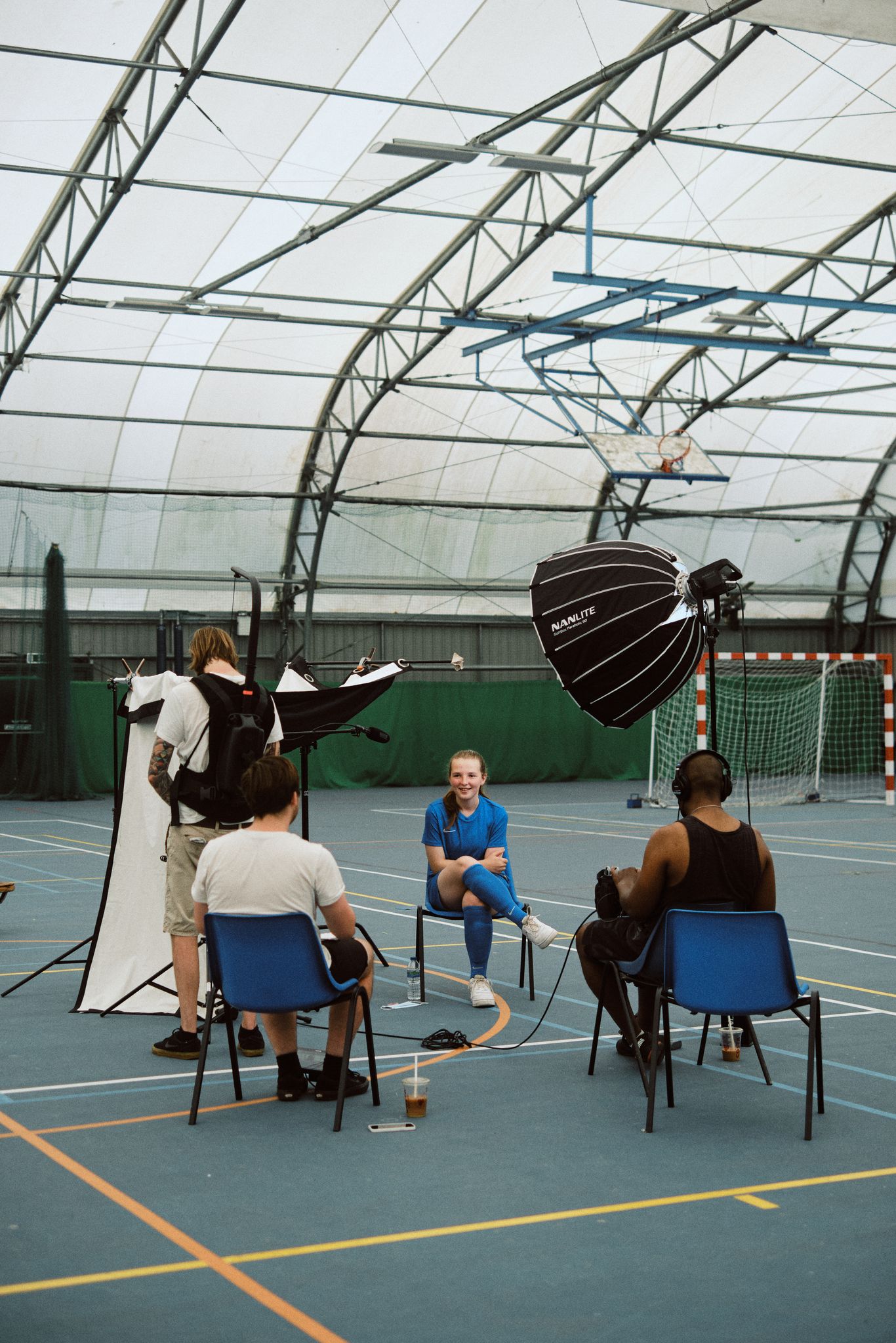 Indoor sports hall setup with tripod, lights, and camera crew surrounding a smiling person in sports attire on a chair, suggesting a relaxed interview.
