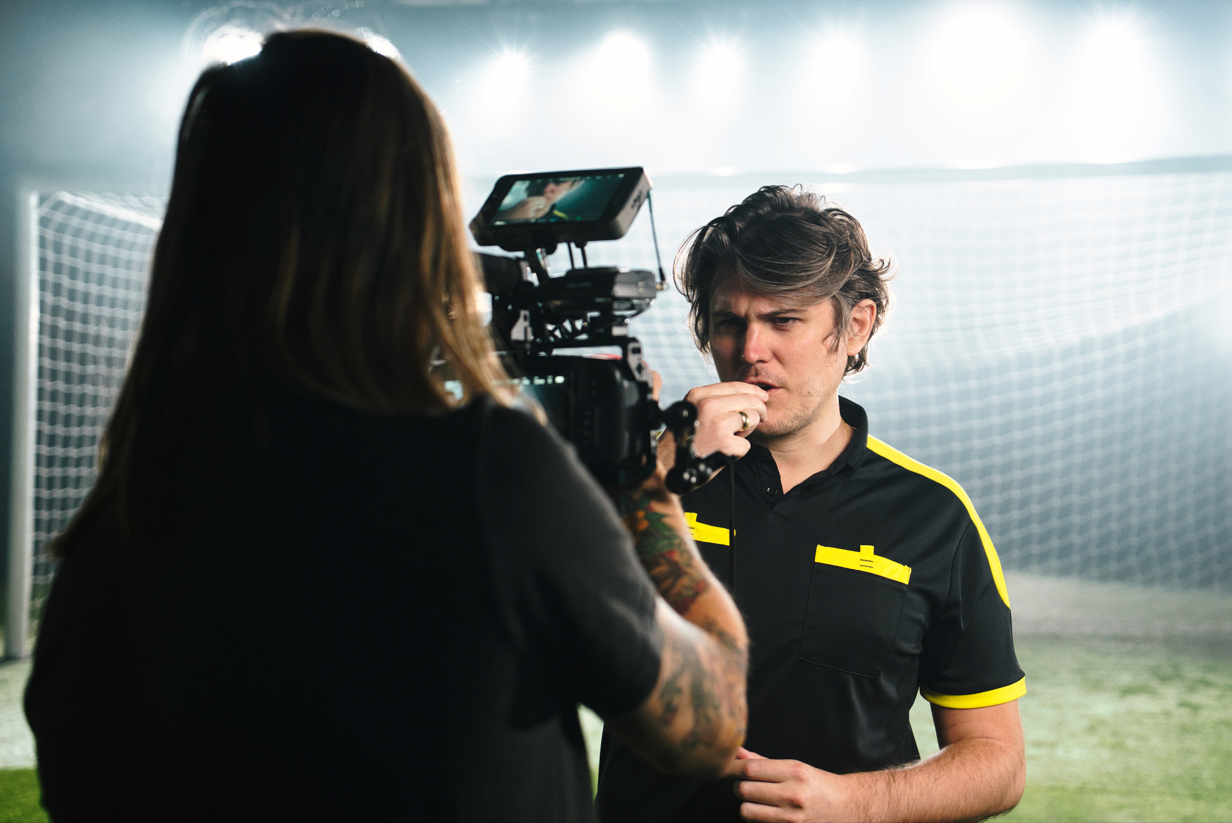 A man in a black and yellow referee shirt is being filmed on a soccer field. He holds a whistle, appearing focused. Bright lights illuminate the scene.
