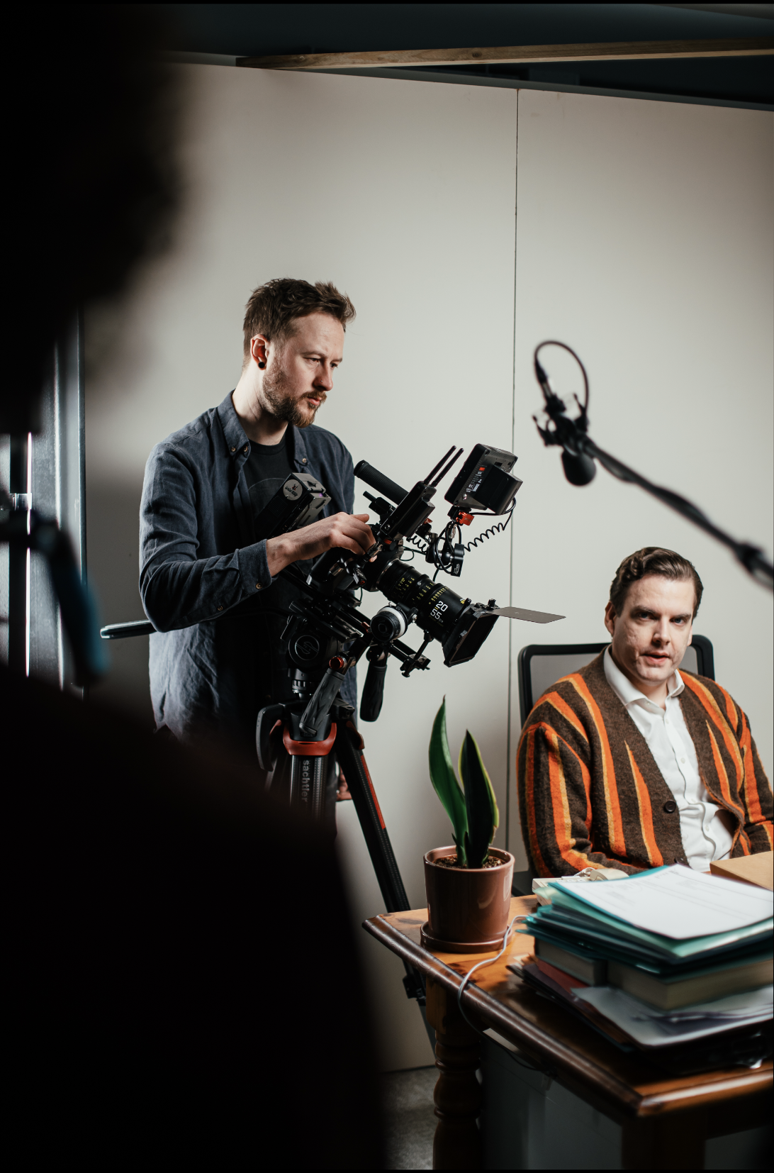 A cameraman operates professional equipment while filming a man in a striped sweater seated at a desk with stacked papers and a potted plant, conveying focus.