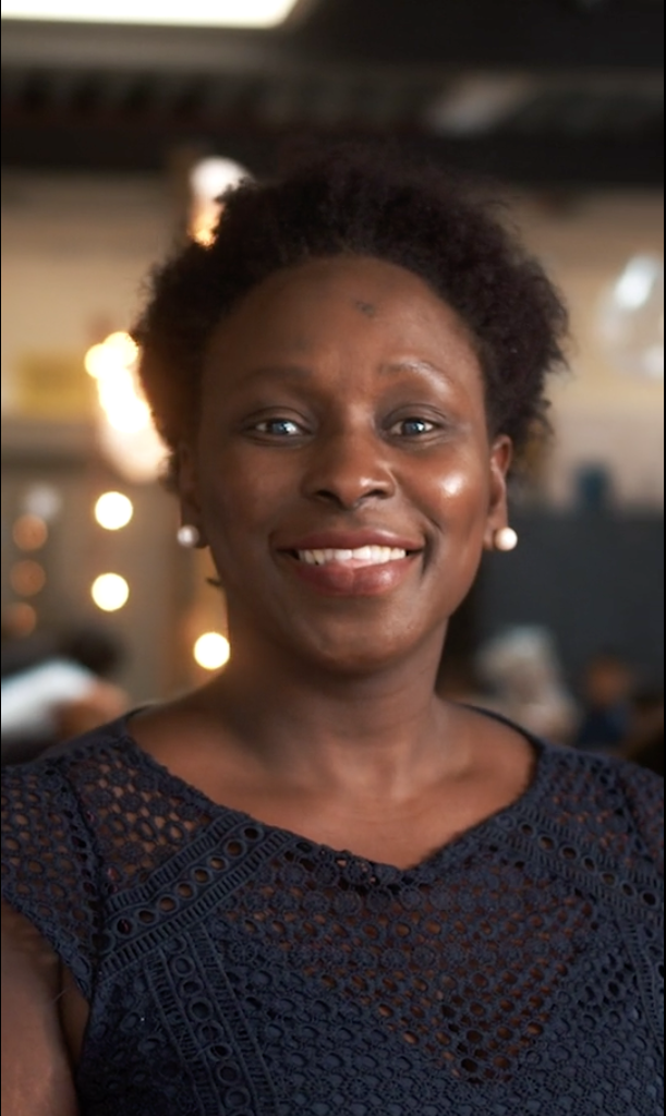 Smiling woman in a dark blue lace dress with pearl earrings stands against a softly lit background, conveying warmth and friendliness.