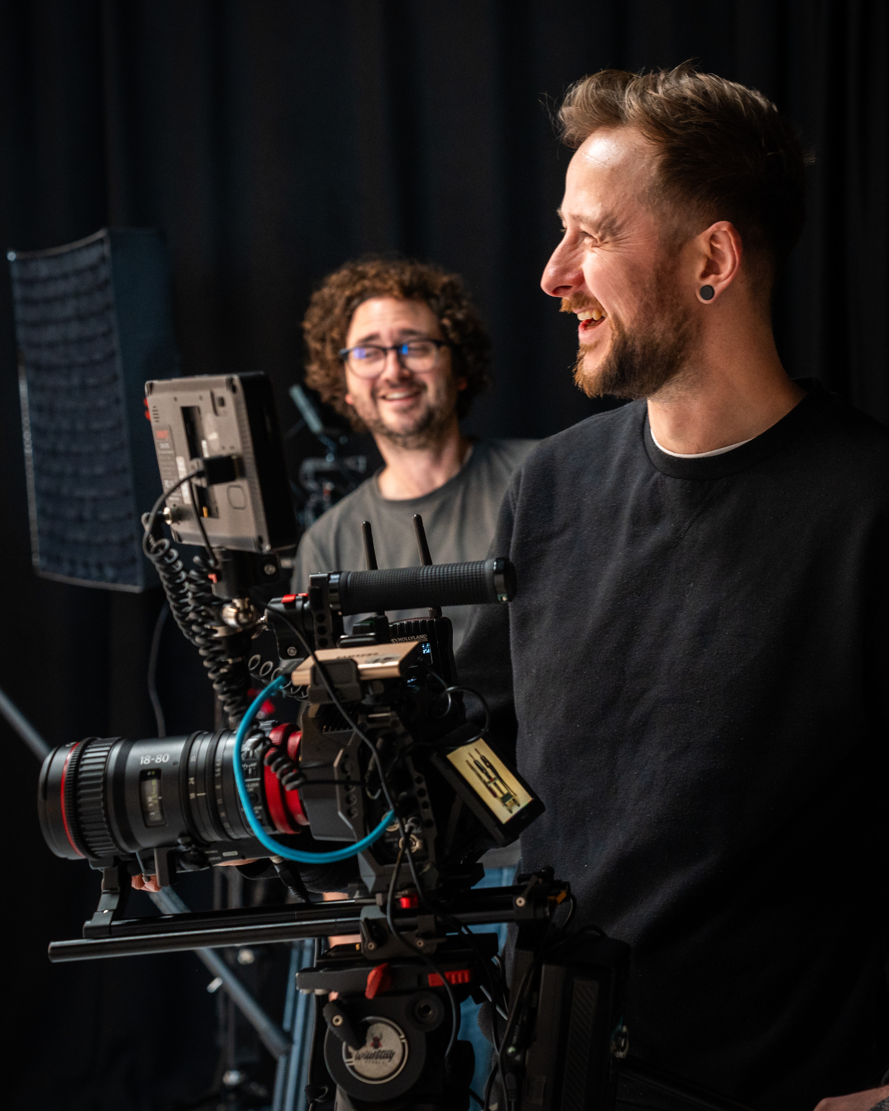 Two men smiling on a film set, one holding a professional camera with a matte box. A studio light is visible in the background. The mood is cheerful.