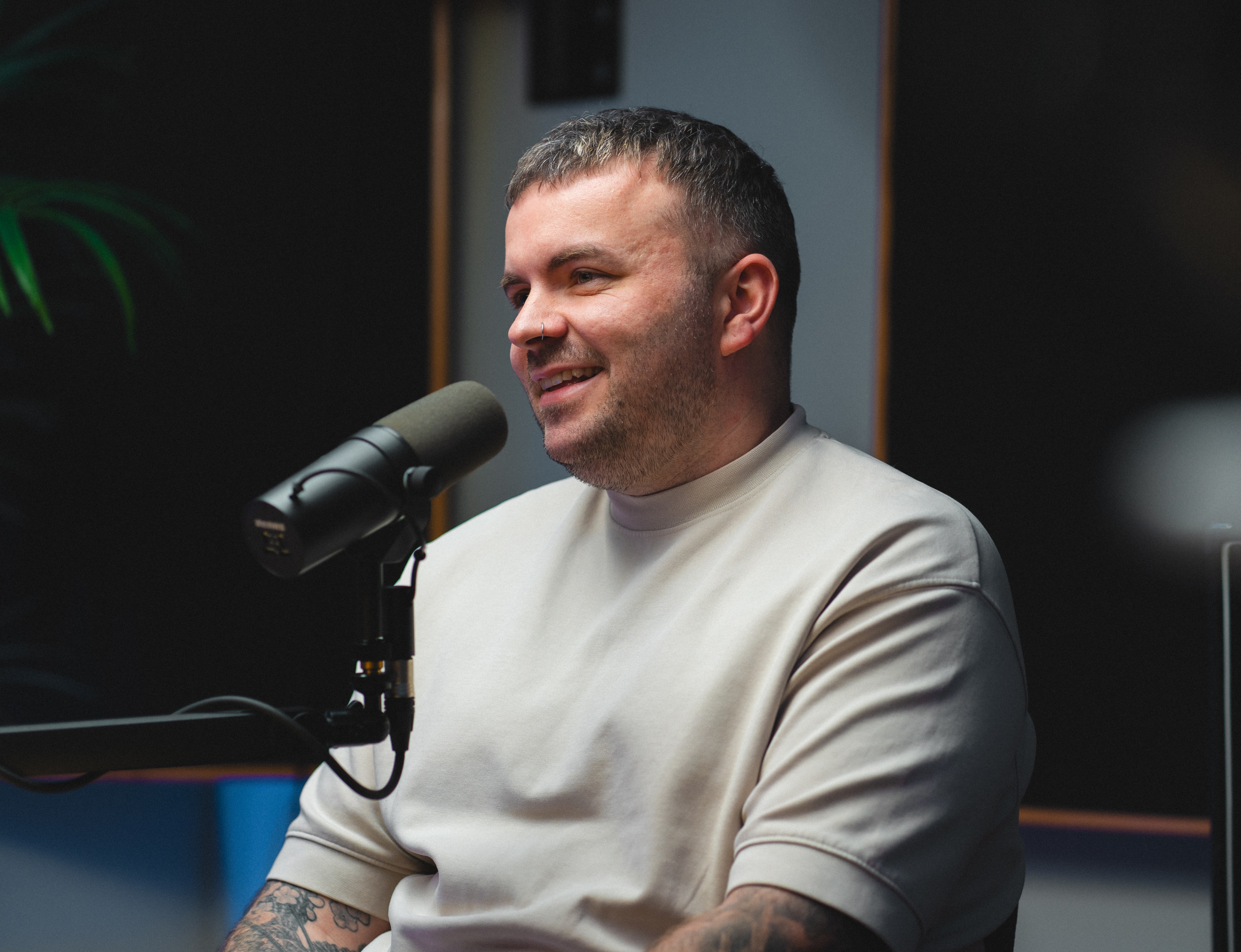 A man with short hair and tattoos sits at a microphone, smiling. He wears a light shirt. The setting appears casual and conversational.