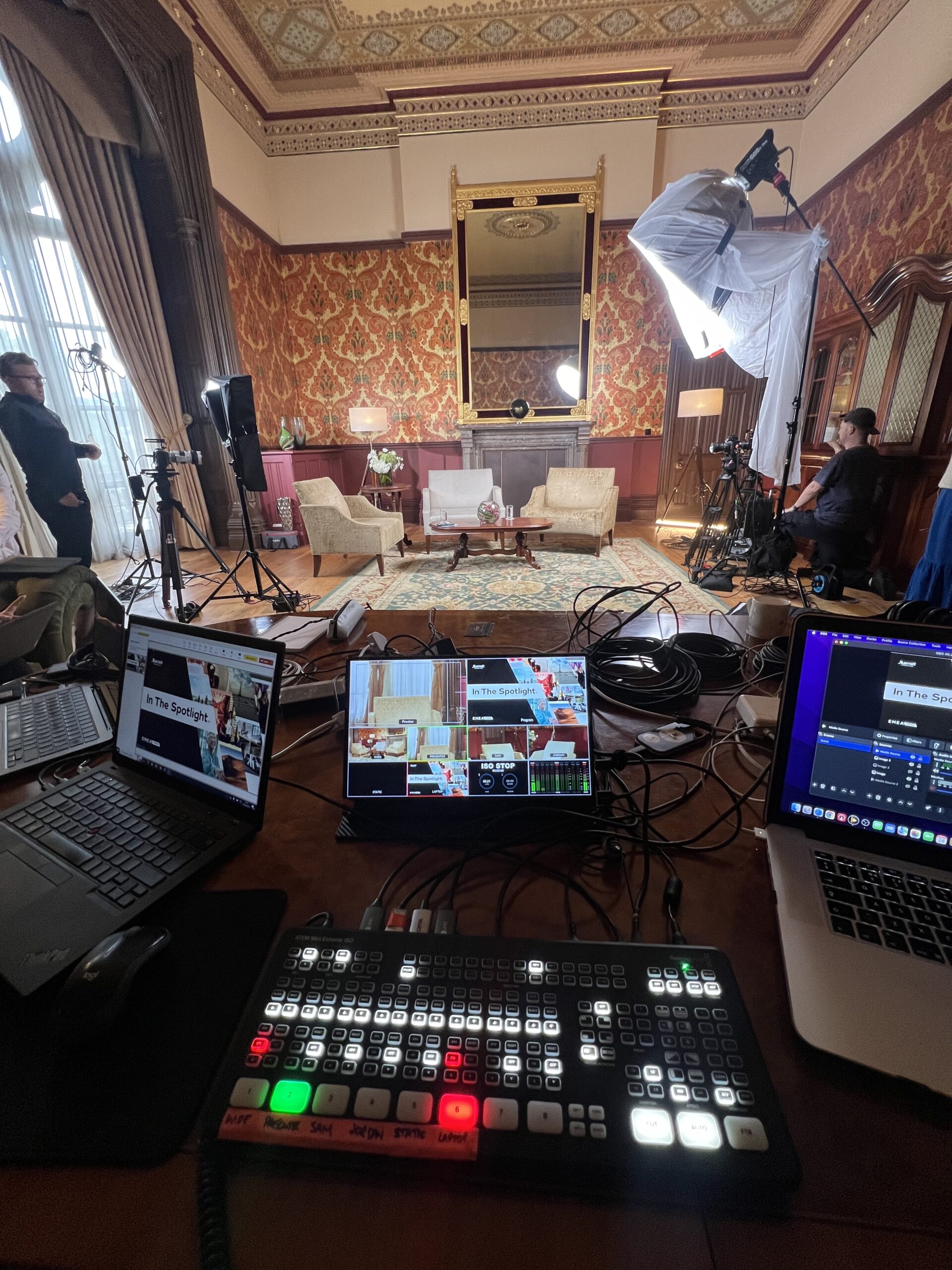 Filming setup in an ornate room with red patterned walls and large mirror. Equipment includes lights, cameras, laptops, and a control panel, creating a professional, focused atmosphere.