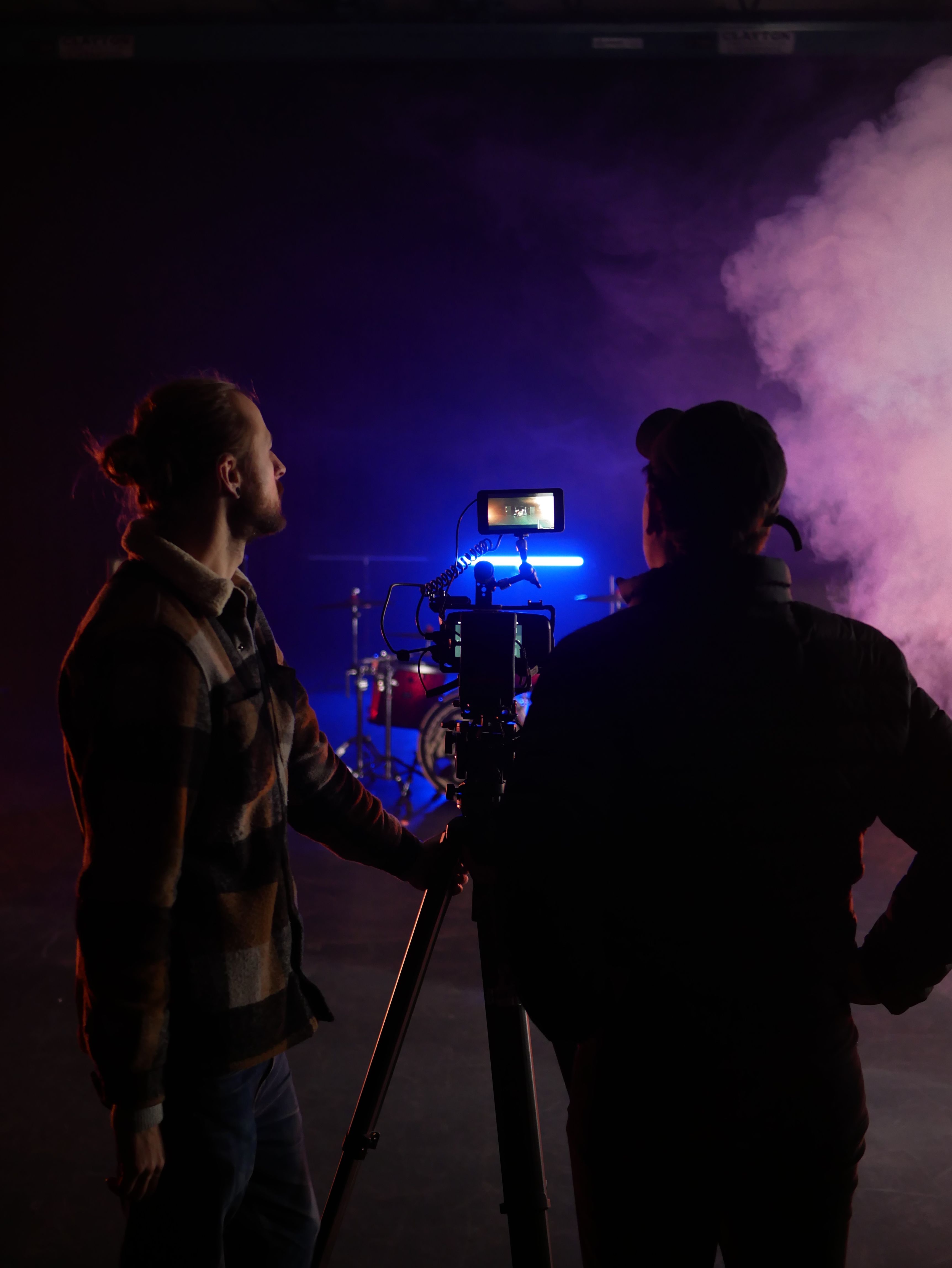 Two people in silhouette stand by a camera tripod in a dimly lit studio. Blue light and fog create an atmospheric scene.