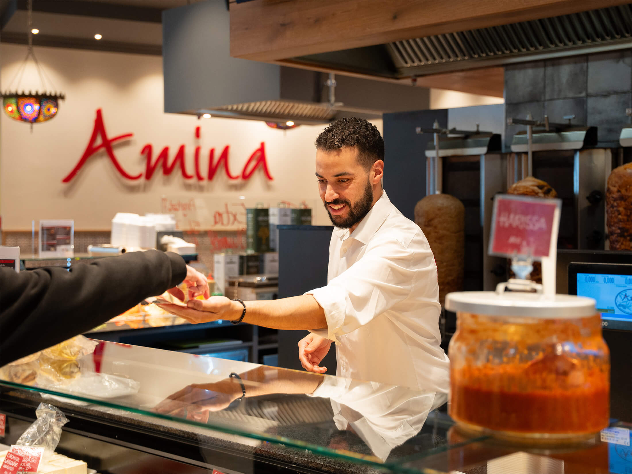 Freundlicher Mitarbeiter begrüßt einen Kunden an der Theke im Amina Feinkostgeschäft.