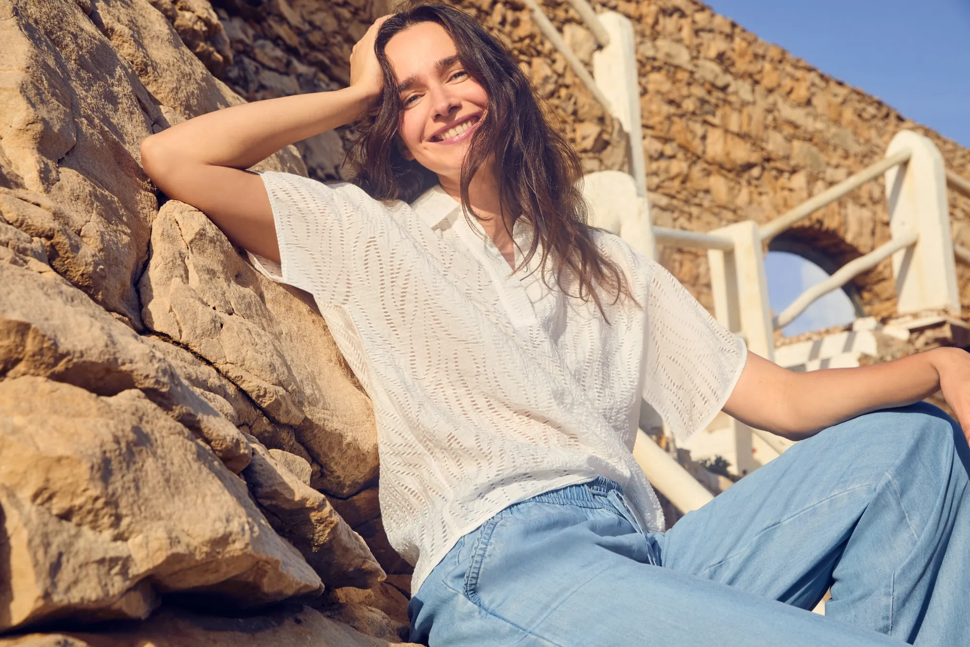 Frau in luftiger weißer Bluse und weiter Jeans sitzend auf Steintreppe – Cecil Sommermode
