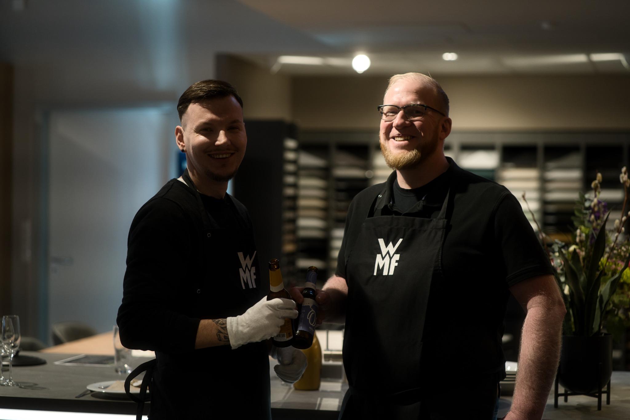 Zwei freundliche Mitarbeiter in WMF-Schürzen stoßen mit Bierflaschen an, aufgenommen in einem modernen Küchenstudio.