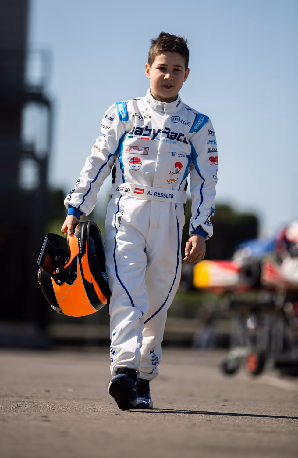 Alexander Ressler spaziert stolz in seinem Rennanzug über das Renngelände. In der rechten Hand hält er seinen Helm.