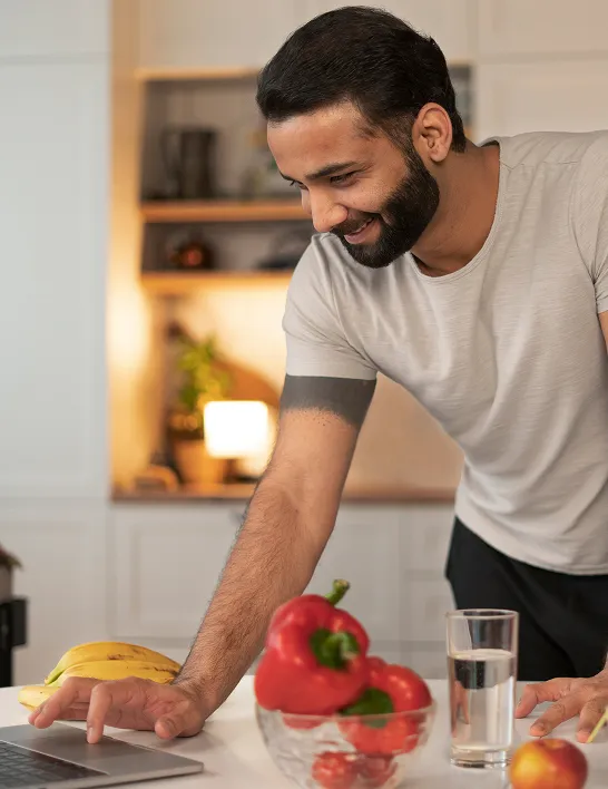 Smiling man using laptop in kitchen with fresh fruits and vegetables, representing healthy lifestyle supported by Erectile Dysfunction Treatment.