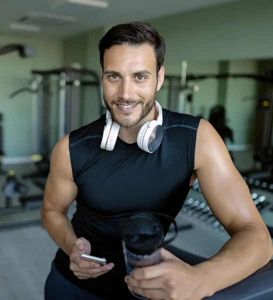 Confident man in athletic wear holding a shaker bottle and phone at the gym, representing improved vitality after GAINSWave® therapy.