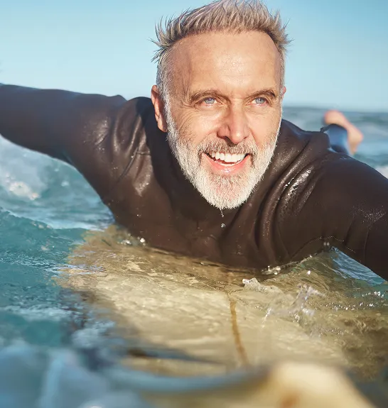 Active older man paddling on a surfboard with a smile, representing enhanced endurance, circulation, and vitality supported by Tri-Amino Acid injections.