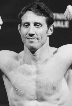 Black and white image of former UFC fighter Tim Kennedy posing shirtless with flexed arms at a weigh-in event, featured as a Gameday Men's Health influencer.