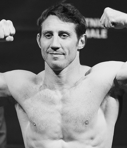 Black and white image of former UFC fighter Tim Kennedy posing shirtless with flexed arms at a weigh-in event, featured as a Gameday Men's Health influencer.