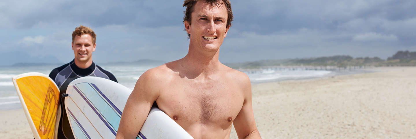 Two men holding surfboards and smiling on the beach, representing active lifestyle benefits associated with P-Shot therapy.