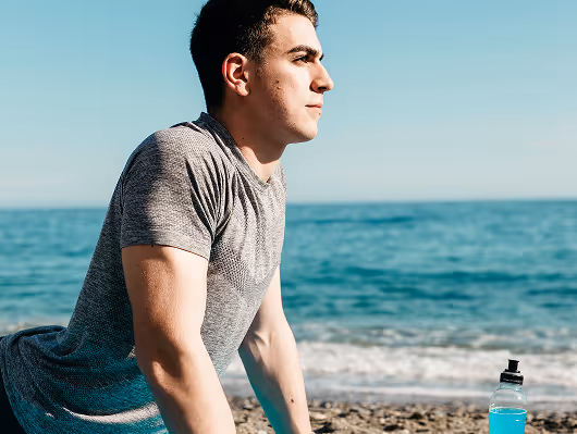 Man in a grey athletic shirt stretching by the ocean with a sports drink bottle nearby, symbolizing recovery and vitality benefits from P-Shot therapy.