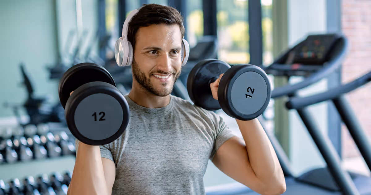 Man smiling while lifting dumbbells in the gym with headphones on, representing fat metabolism and increased energy from MIC lipotropic injections.