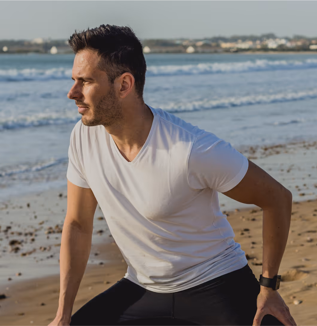 Fit middle-aged man performing a dynamic stretch on the beach, representing improved circulation and performance benefits of GainsWave therapy.