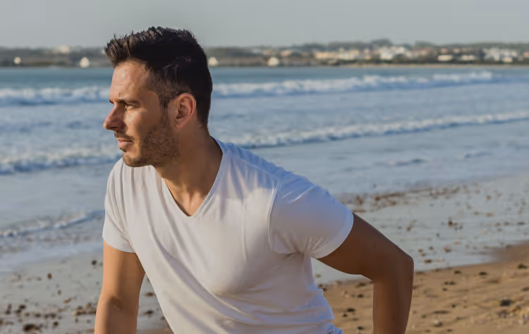 Fit middle-aged man performing a dynamic stretch on the beach, representing improved circulation and performance benefits of GainsWave therapy.