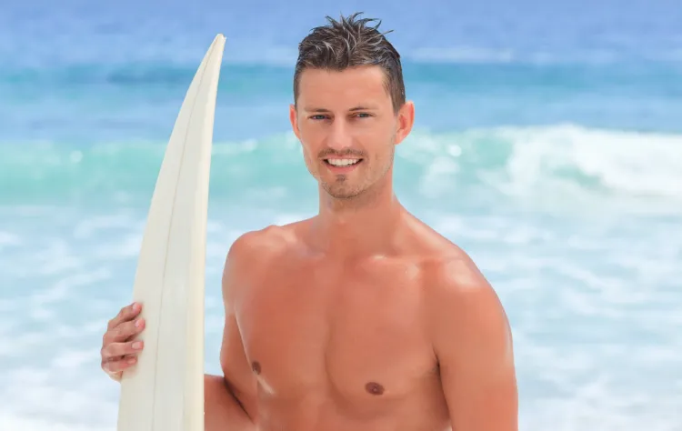 Fit man standing on a beach holding a surfboard, smiling with the ocean waves in the background.