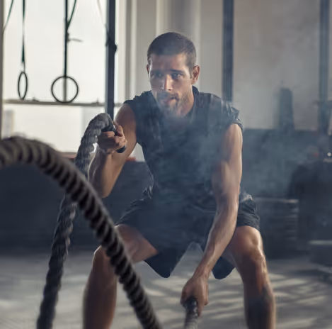 A man in athletic clothing exercises with battle ropes in a gym, focusing on fitness and mens weight loss with phentermine, with gym equipment and light streaming in the background.