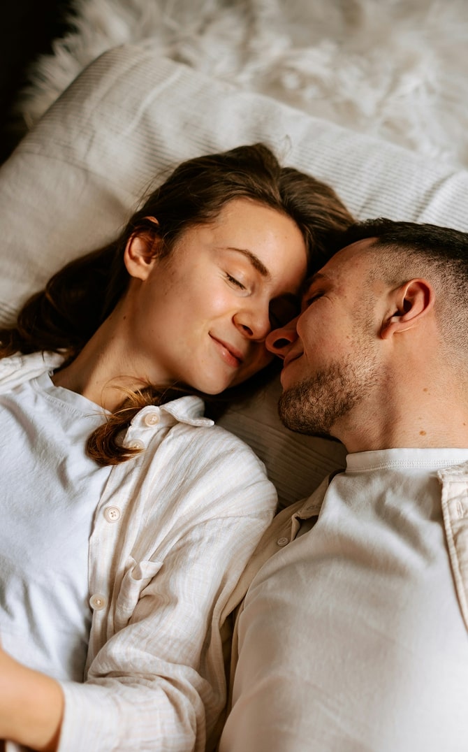 Young couple laying next to each other in bed, smiling with their eyes closed.