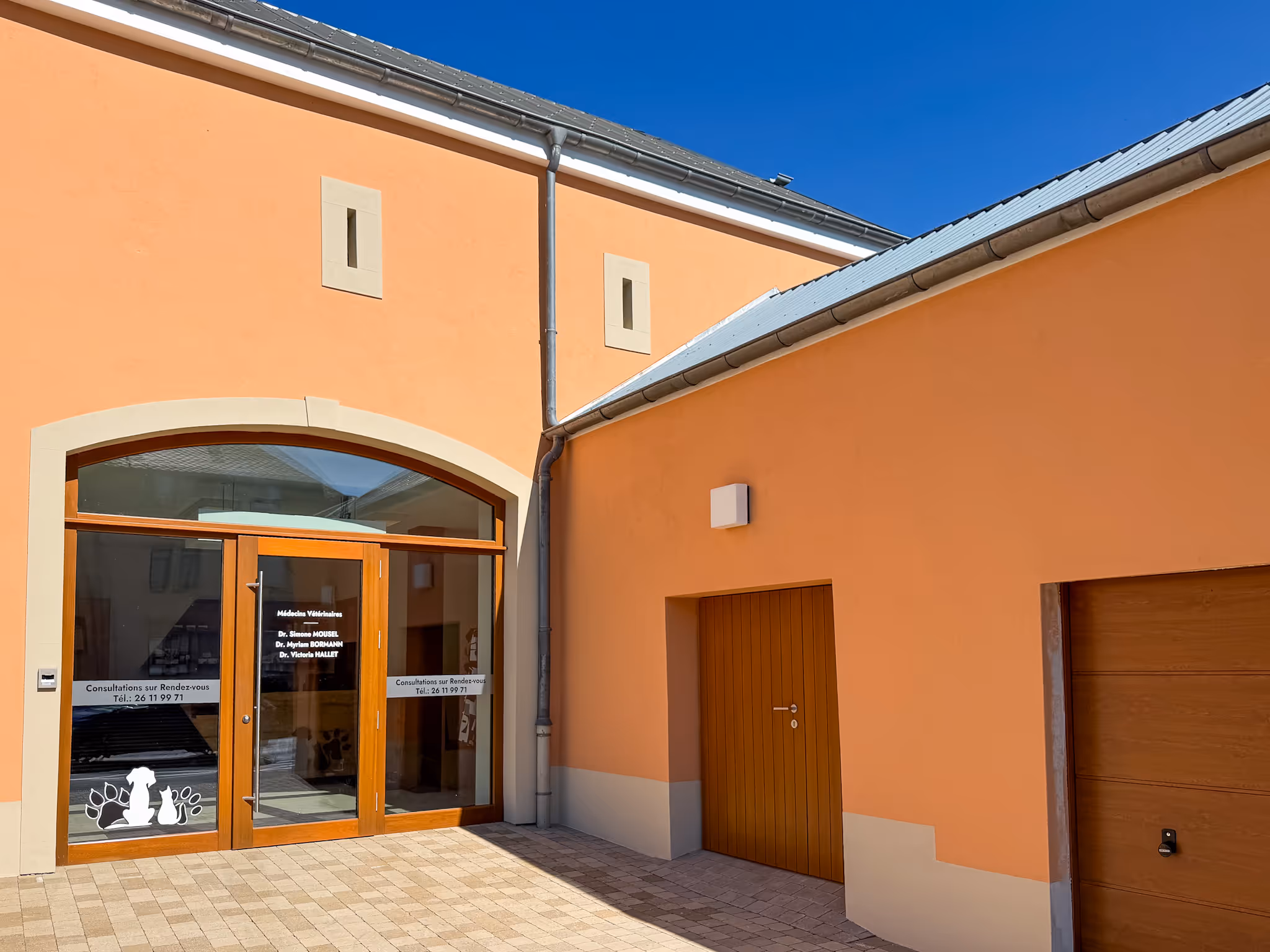 Orange building with small windows, an arched doorway, and a paved courtyard under a clear blue sky.