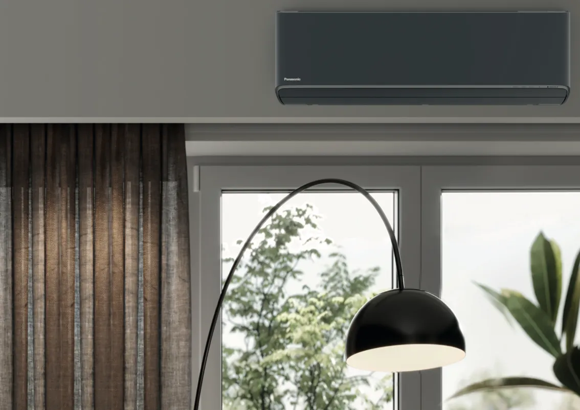 Een antracietkleurige Panasonic lucht-luchtwarmtepomp aan de muur van een woonkamer, naast een raam met gordijnen en een booglamp.