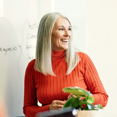 Smiling woman with long gray hair wearing an orange ribbed turtleneck sitting near a whiteboard.