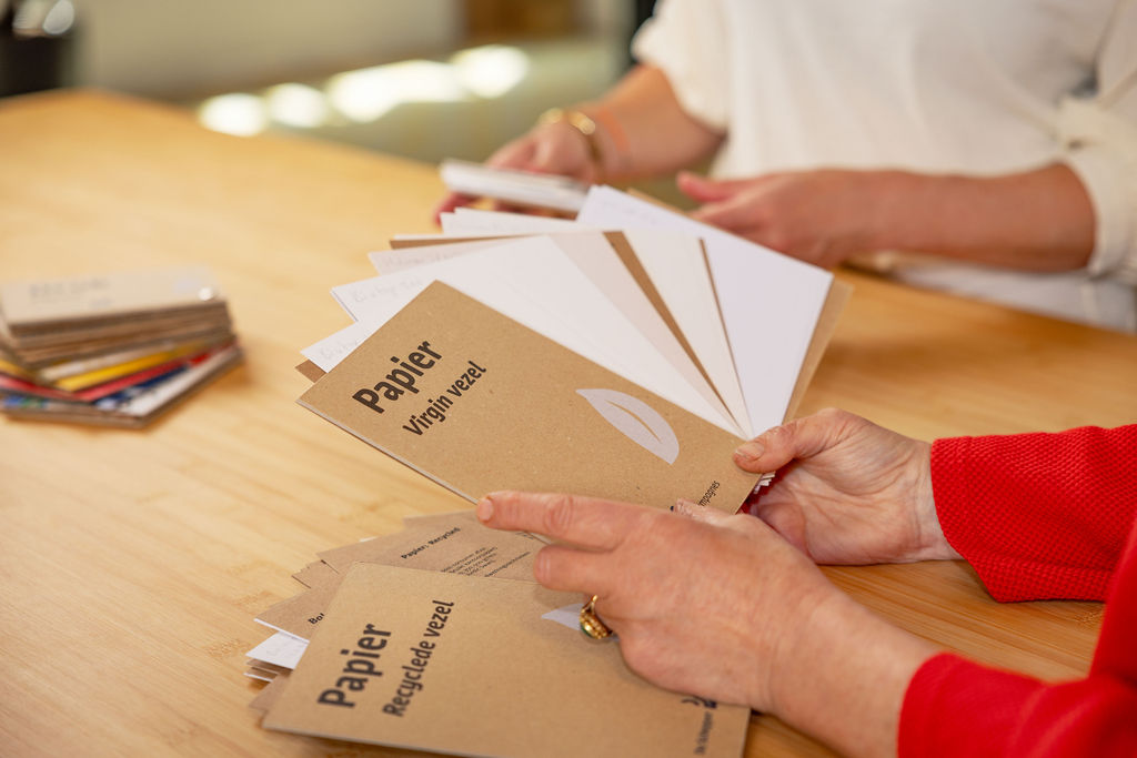Hands holding a fan of paper sample swatches labeled with types of paper fibers on a wooden table.