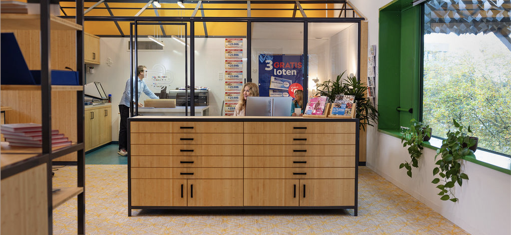 Modern office reception with wooden desk, two women seated behind computers, man at a printer in the background, and large windows with plants.