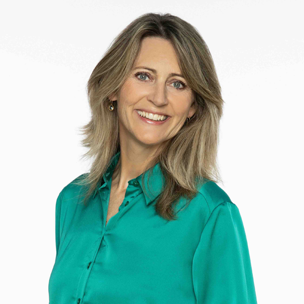 Smiling woman with shoulder-length blonde hair wearing a teal button-up shirt against a white background.