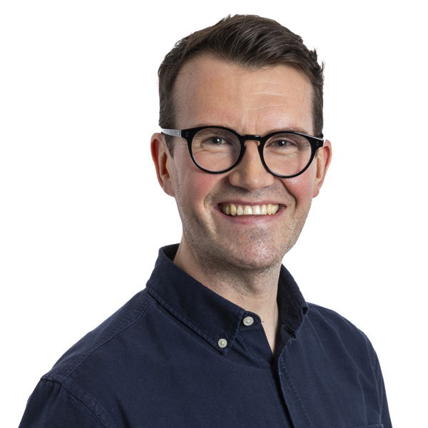 Smiling man with short brown hair and round black glasses wearing a dark blue button-up shirt.
