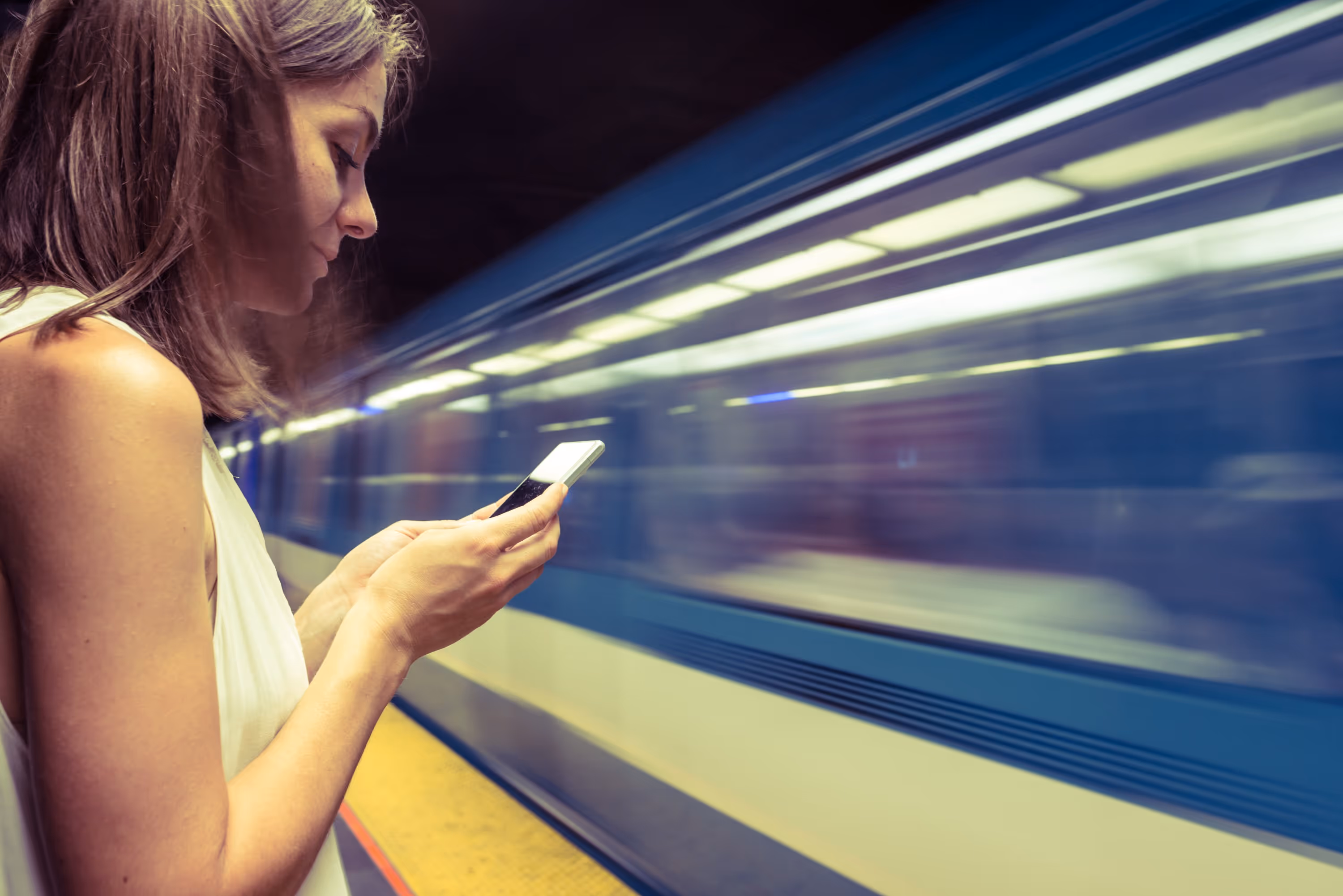 Femme utilisant son smartphone sur un quai de métro, un train en mouvement en arrière-plan, illustrant l'intégration technologique dans la mobilité.
