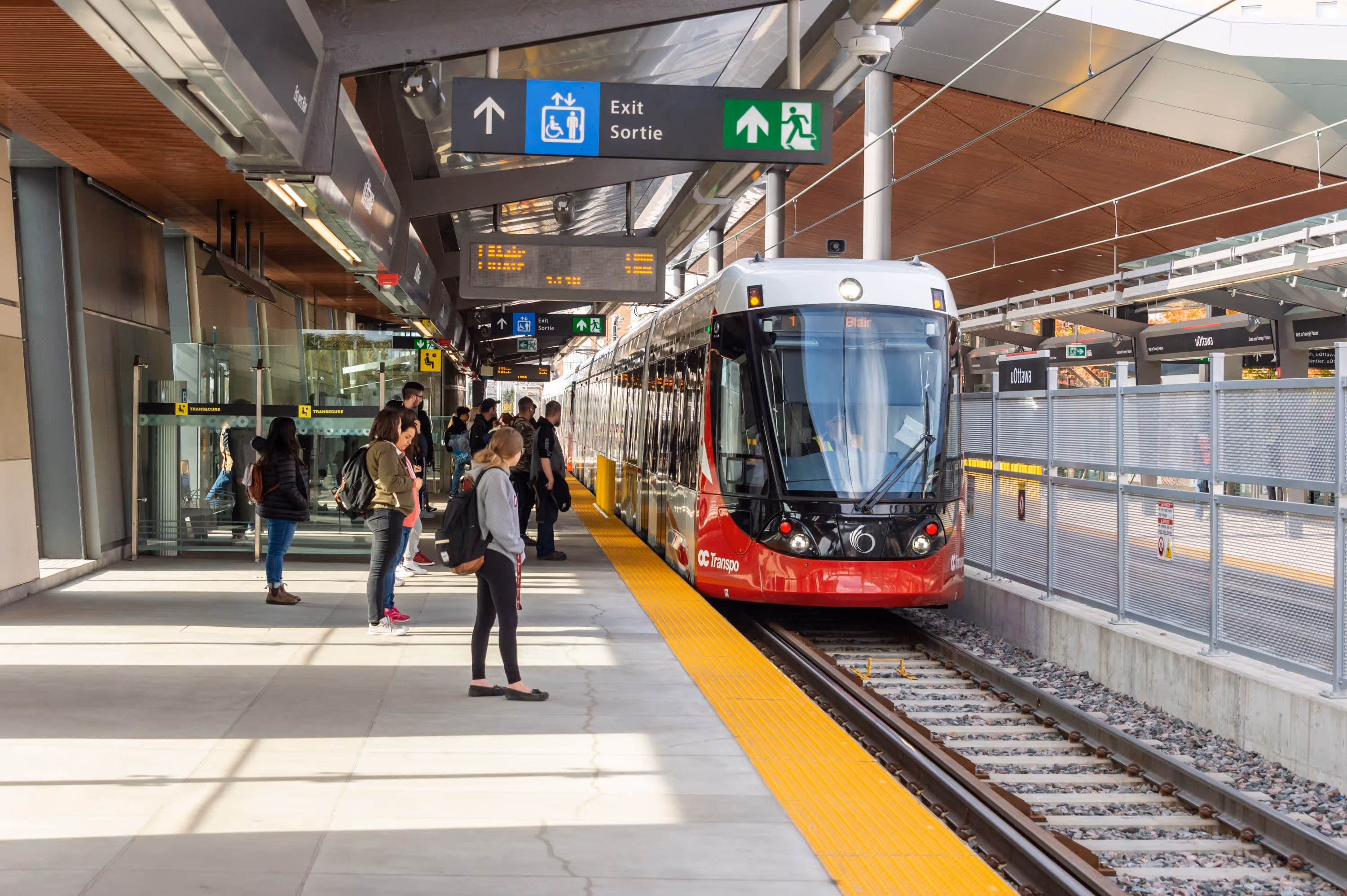 Train léger d'OC Transpo à une station moderne à Ottawa, passagers sur le quai, signalisation de sortie claire.