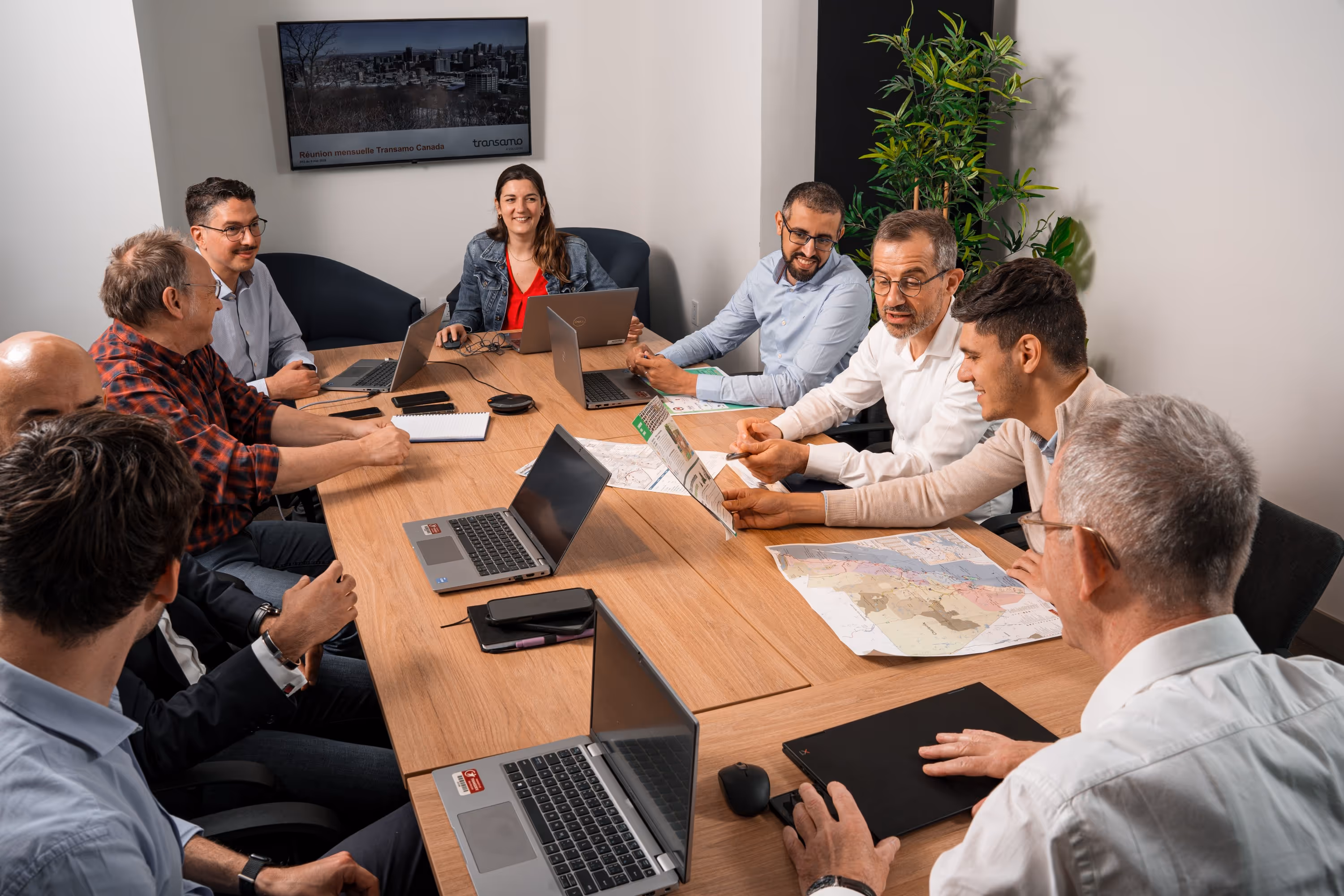 Réunion d'équipe complète de Transamo Canada, des professionnels discutant et collaborant autour d'une table avec cartes et ordinateurs portables.