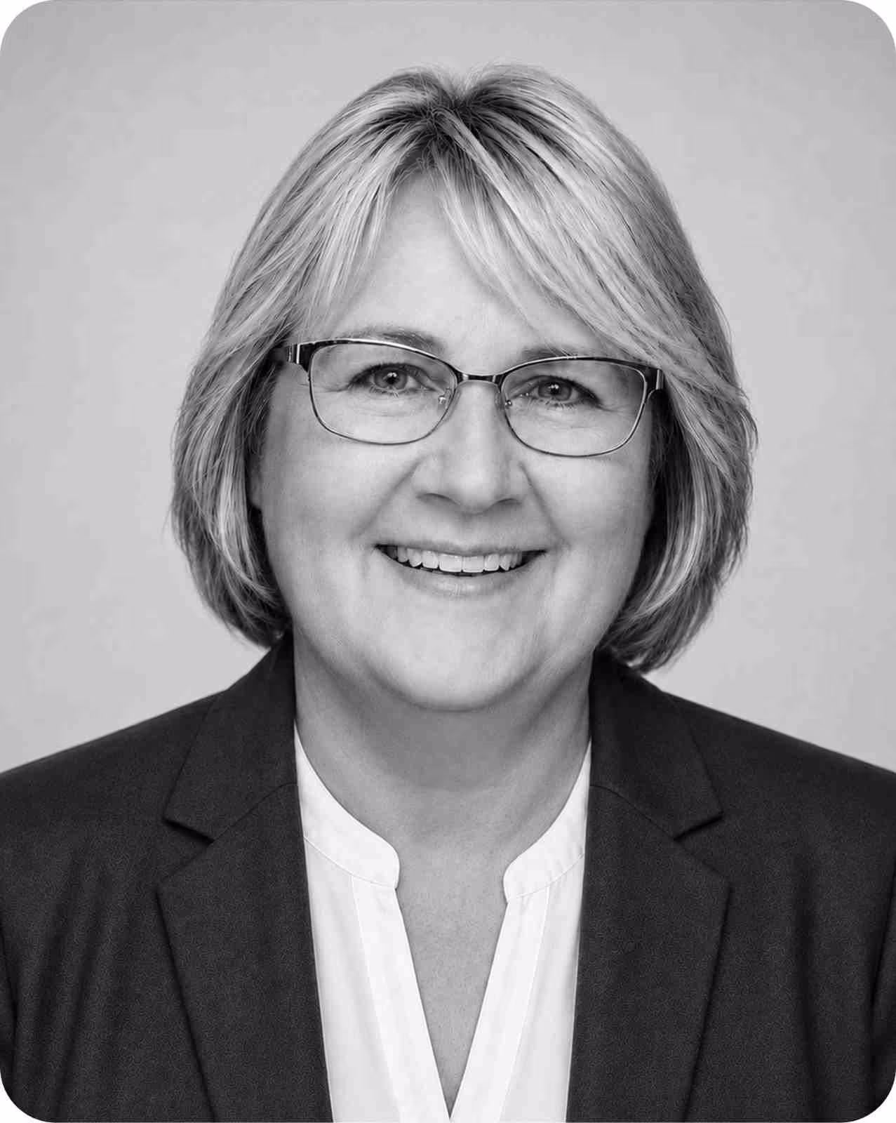 Smiling middle-aged woman with glasses and short light hair wearing a blazer and blouse.