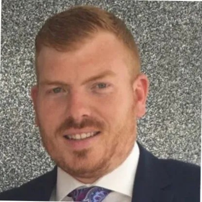 Portrait of a smiling man with short red hair and a beard, wearing a suit and paisley tie, against a speckled gray background.