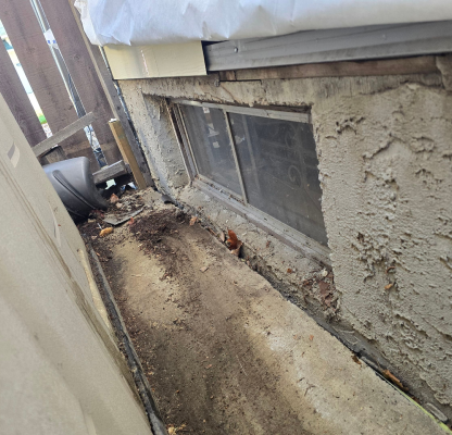 Narrow side yard with a dirty concrete pathway, a basement window with metal bars, and weathered stucco wall.