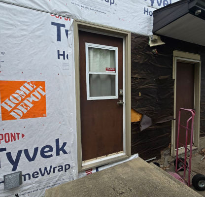 Brown door with a frosted window and a 'Falco' sticker, surrounded by black and white house wrap with a Home Depot logo, next to a dolly on concrete steps.