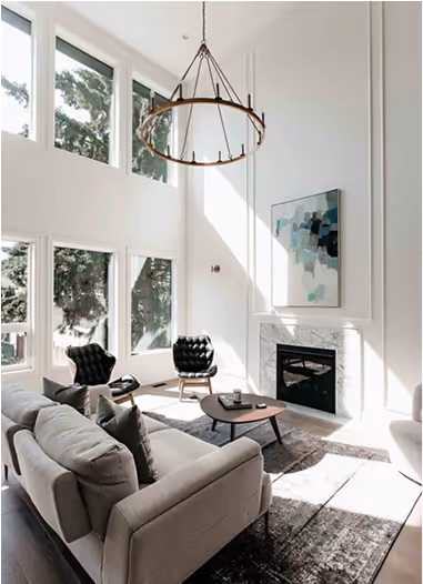 Bright living room with tall windows, a modern chandelier, light gray sofa, two black chairs, a coffee table, and a fireplace with abstract painting above it.