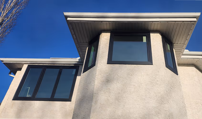 View looking up at the corner of a beige stucco house with multiple black-framed windows under a clear blue sky.