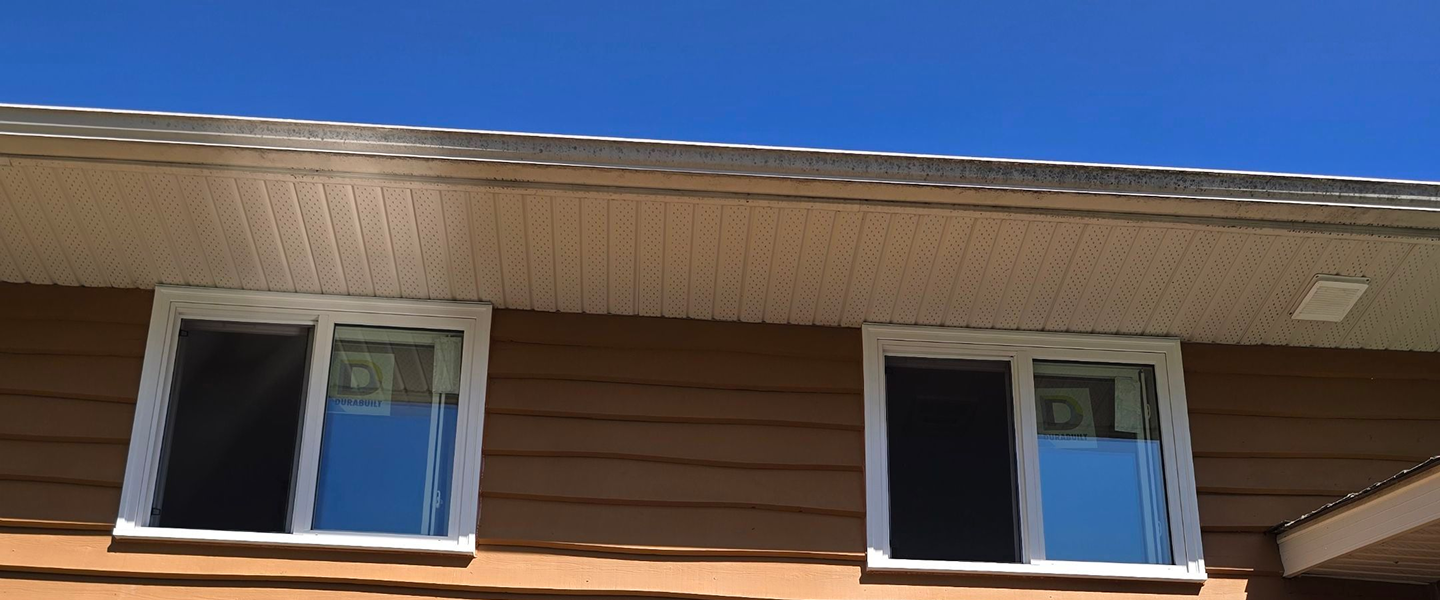 Two adjacent double-pane windows with white frames on a brown house exterior under a white soffit and clear blue sky.