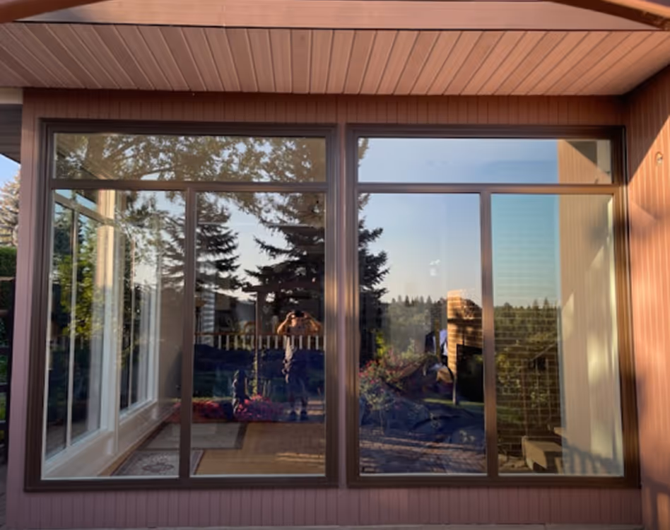 Large glass sliding door with brown frame reflecting outdoor trees, patio, and a person taking a photo.