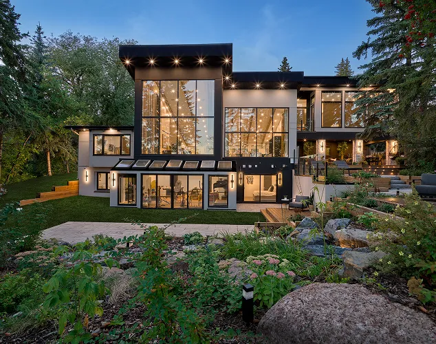 Modern two-story house with large floor-to-ceiling windows illuminated at dusk, surrounded by lush greenery and a landscaped garden.