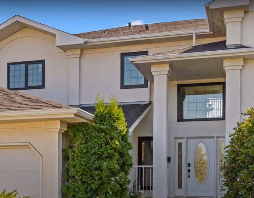 Modern two-story house with beige exterior, large windows, and a front glass door surrounded by green shrubs.