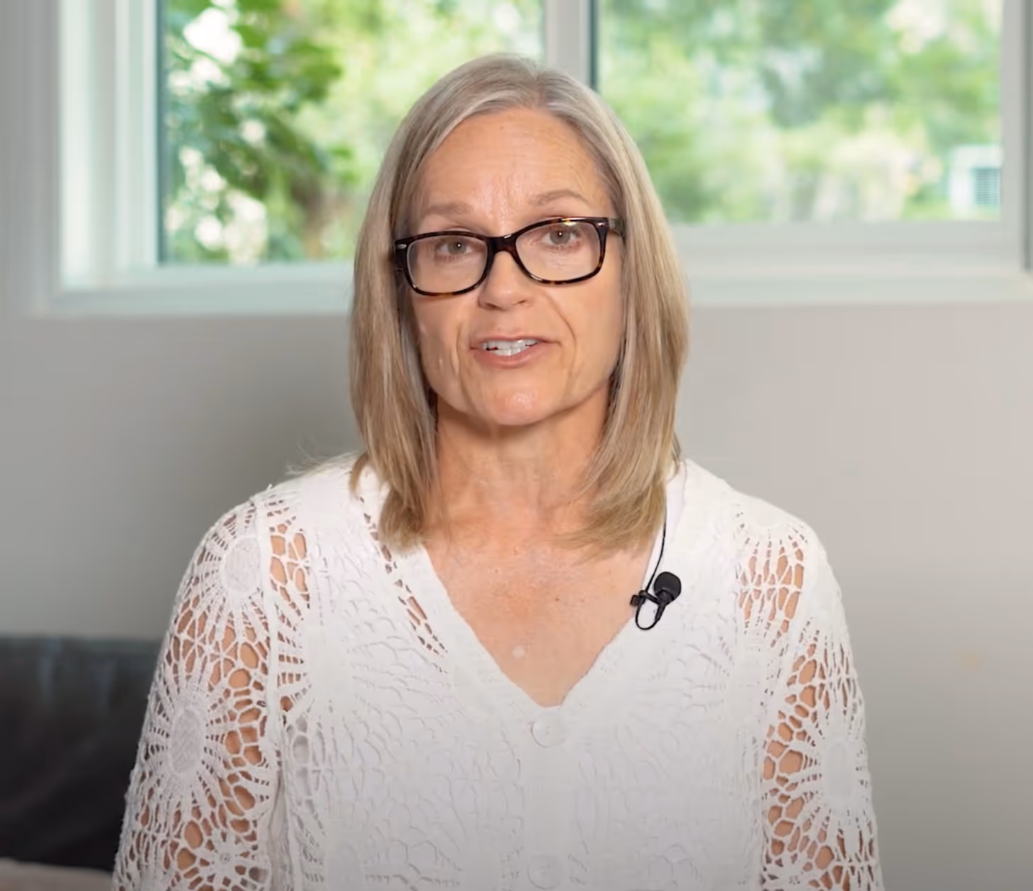 Middle-aged woman with glasses wearing a white crocheted top speaking indoors with a window and greenery in the background.