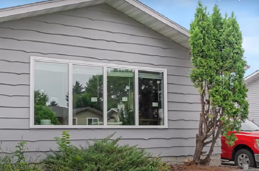 Side view of a gray house with horizontal siding, a large window reflecting greenery, and a tall bush next to a red truck.