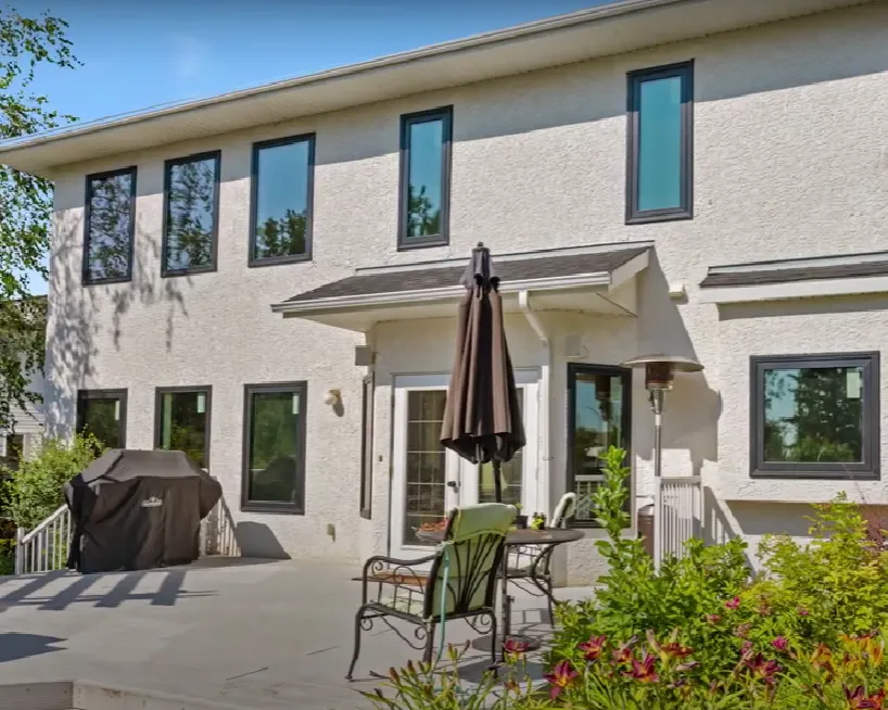 Backyard patio of a two-story house with multiple tall windows, patio furniture, a closed umbrella, a grill, and greenery in the foreground.